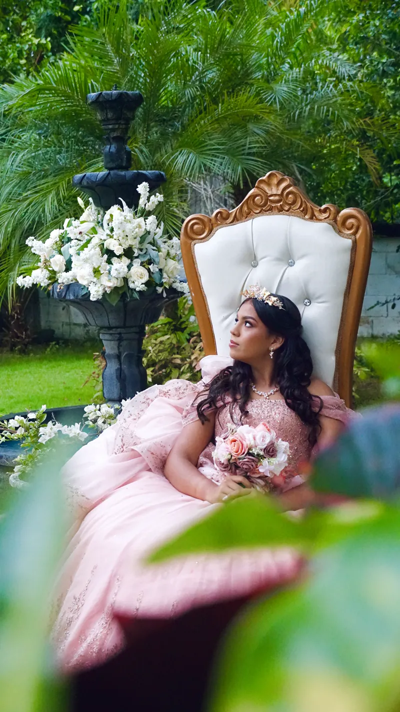 Quinceañera con corona en un sillón blanco y dorado, junto a una fuente con flores blancas